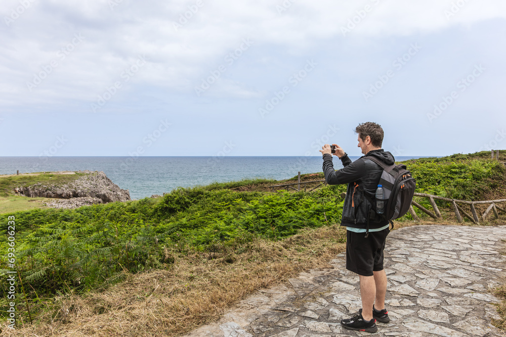 Middle-aged man with backpack and casual clothes taking a photo with his mobile phone on a path by the sea. Tourism and travel concept