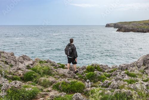 Back view of a mature man with backpack and casual clothes looking at the sea with his dog in nature. Travel and nature concept