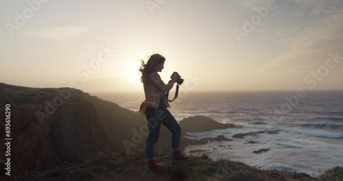 Epic and cinematic shot of female adventure or travel photographer stand on edge of cliff in evening sunset light. Wind blow her hair around. Explore exotic travel destination and create content photo