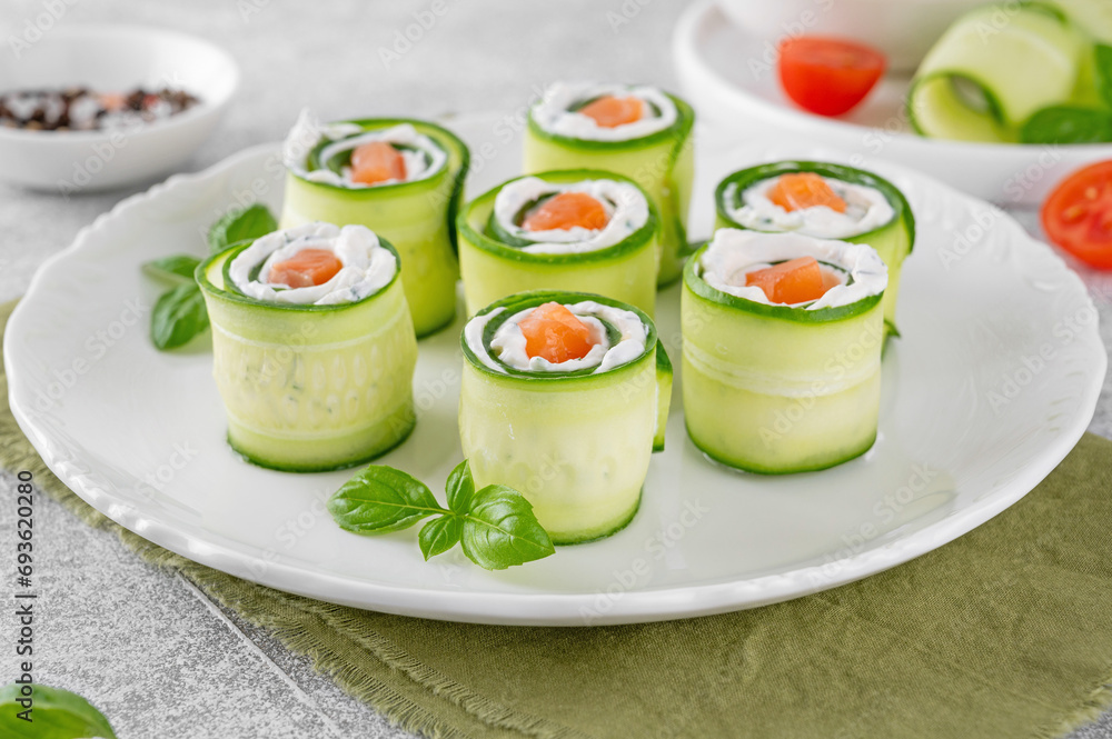 Cucumbers rolls with cream cheese, salted salmon and fresh herbs served on a white plate on a concrete background. Selective focus Cucumbers rolls with cream cheese, salted salmon and fresh herbs served on a white plate on a concrete background. Selective focus