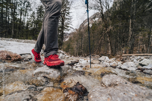 Hiking or trail running in the mountains. Outdoor cross-country running in mountains. Hiking, exercising, fitness and healthy lifestyle. Red running shoes jumping over a creek. Trekking poles.