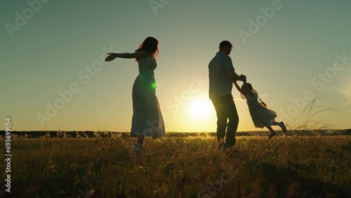 Family enjoys tranquility of park among waving grass forming memories on journey. Mother dances and father spins daughter holding hands at dusk. Family silhouettes in field of swaying grass at sunset