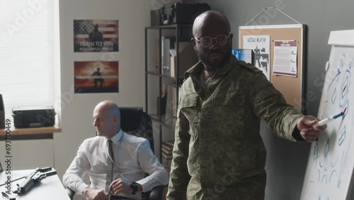 Medium long side shot of African American male cadet standing near whiteboard explaining chosen tactics to Caucasian military professor during class in academy photo