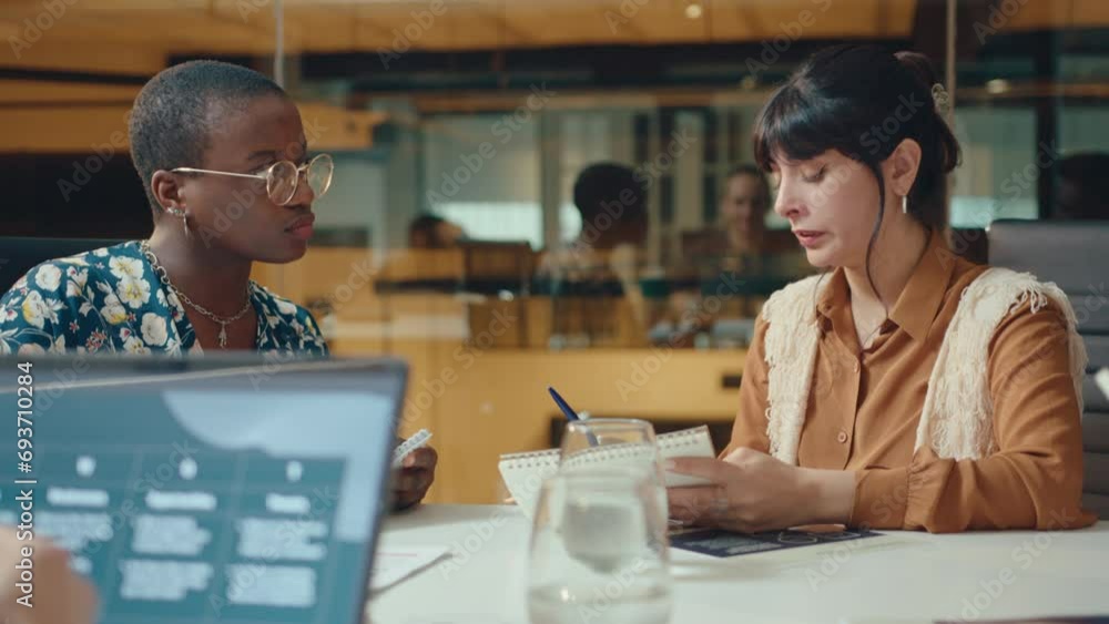 Young Latin businesswoman reading notes in copybook and having conversation with Black female colleague at meeting table in coworking room Young Latin businesswoman reading notes in copybook and having conversation with Black female colleague at meeting table in coworking room
