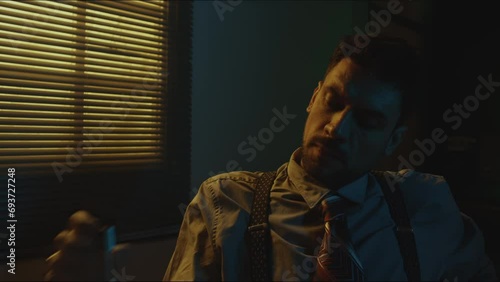 Chest up portrait of Caucasian tired and perplexed man in vintage style officewear playing with lighter, drinking liquor and then looking at camera in dimly lit room photo