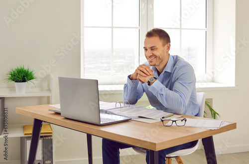 A young attractive businessman working on a laptop at the office with financial documents at the desk with charts and tables. Satisfied confident man chatting online or having video conference call.