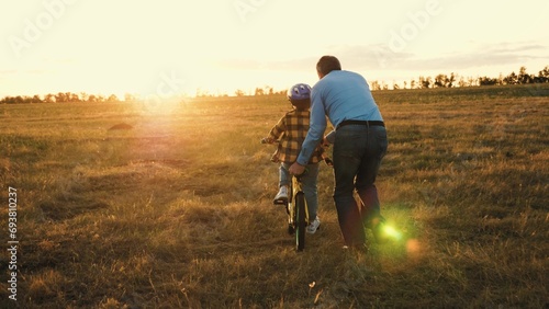 Affectionate papa actively involved in assisting little kid on riding bicycle. Caring dad prevents mishaps holding bike of son. Father provides risk-free bike ride for beloved child in field photo