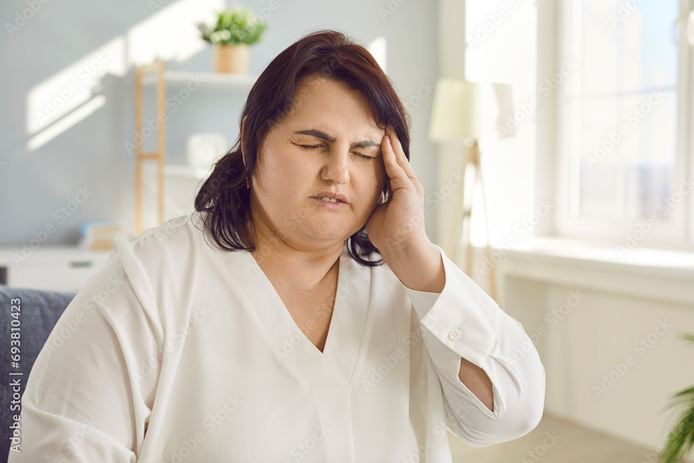 Overweight woman suffering from sudden attack of headache or dizziness. Portrait of exhausted plus size woman sitting on sofa touching her temple feeling fatigue, nervous tension Overweight woman suffering from sudden attack of headache or dizziness. Portrait of exhausted plus size woman sitting on sofa touching her temple feeling fatigue, nervous tension