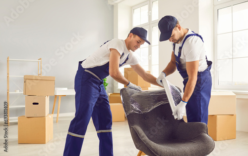 Portrait of a two young smiling male movers packing furniture in the living room. Moving service men workers wrapping chair with plastic wrap. Move, moving day and relocation concept. photo