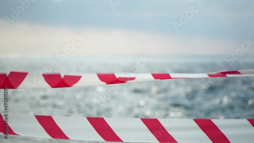 Red white warning tape barrier ribbon swinging in the wind across exotic sea beach background without people. No entry Red White caution tape. No holiday concept, delayed travel, no summer plans photo