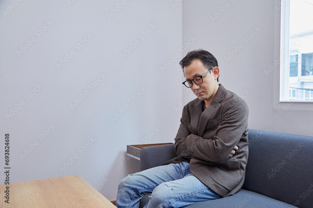 Asian businessman wearing eyeglasses in dark suit, taking a nap on grey sofa in modern living room, tired and exhausted from busy day at workplace.
