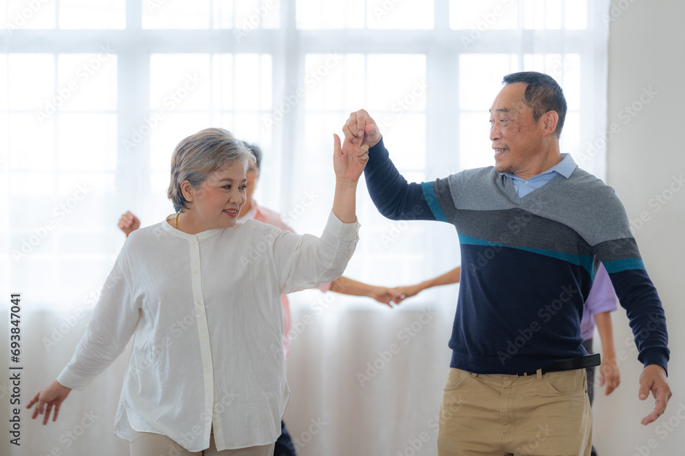 Obraz premium Asian Older male and females people dance with their partners on a dancing floor in living space. Happy older couple performing get exercise. Joyful carefree retired senior friends enjoying relaxation