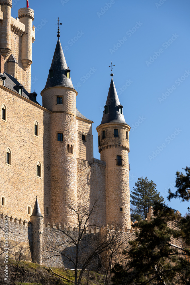 Fototapeta premium Majestic castle of Segovia, Spain with towering spires against a clear blue sky, surrounded by trees.