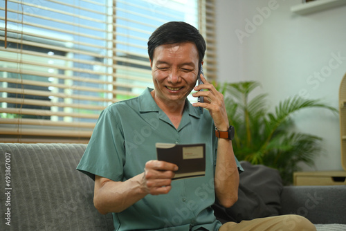 Smiling senior man looking at passbook while talking on mobile phone with customer support operator.