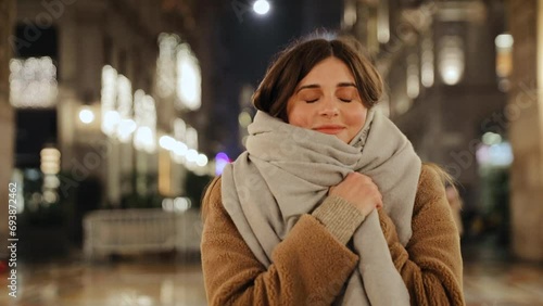 Woman covers her body with a scarf and close her eyes. Woman wearing warm clothing. Steadicam shot on the night street of Milan, Italy photo