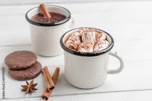 Homemade spicy hot chocolate drink with marshmallows in enamel cup on white wooden table with cinnamon stick, cookies and star anise