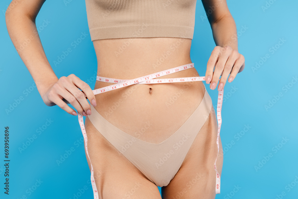 Attractive thin woman in underwear measuring waist with measure tape. Isolated on blue background. Fitness, diet, weight loss concept