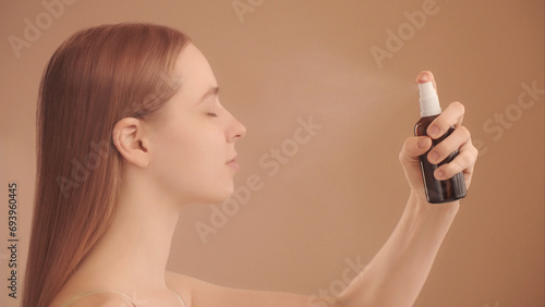 Young woman sprays a facial spray mist or thermal water, soothing tonic. Side view, spraying process, water droplets flying. Toning, moisturizing, soothing facial skin. Peach Background photo