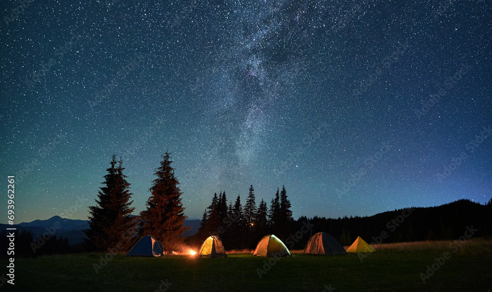 Night camping in mountains under starry sky. Tourist tents in campsite near burning campfire under beautiful sky full of stars with Milky way above forest. Concept of tourism and traveling. - obrazy, fototapety, plakaty 