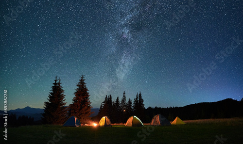 Night camping in mountains under starry sky. Tourist tents in campsite near burning campfire under beautiful sky full of stars with Milky way above forest. Concept of tourism and traveling. photo