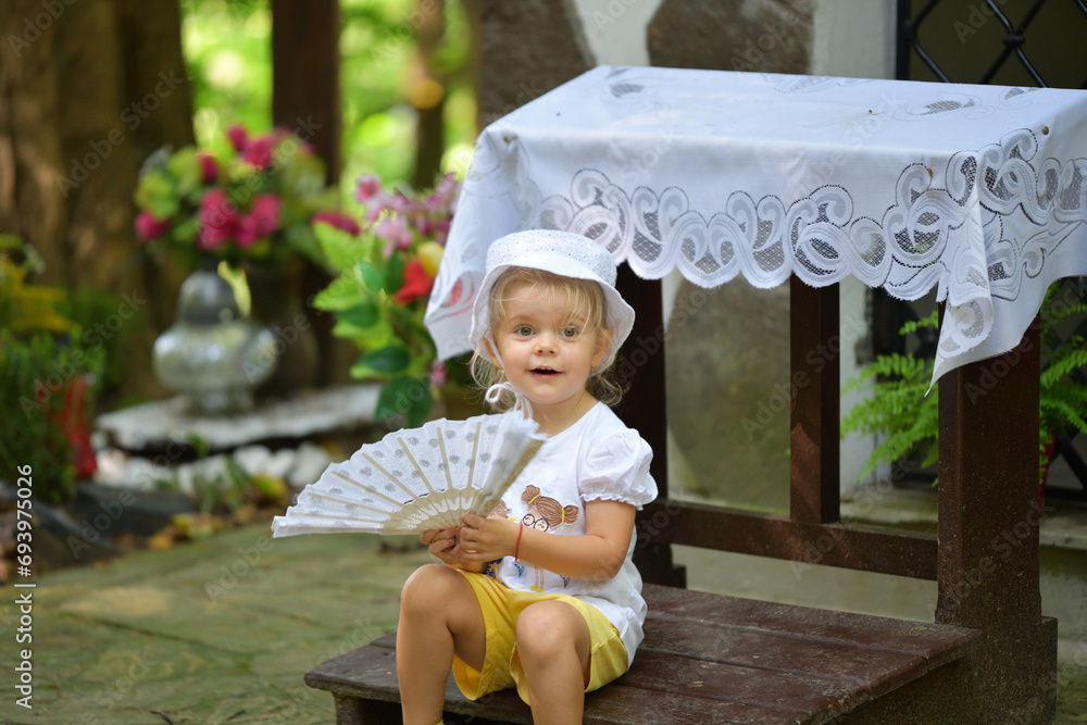 The little girl sits by the forest chapel and waves her hand with a fan like a princess