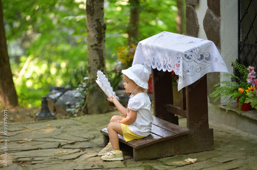 A little girl is sitting in the forest and waving a cloth fan in front of her to cool off