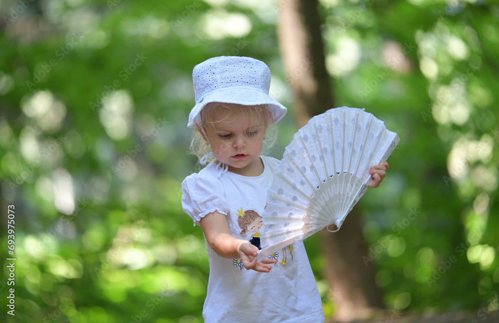 A little girl is sitting in the forest and waving a cloth fan in front of her to cool off