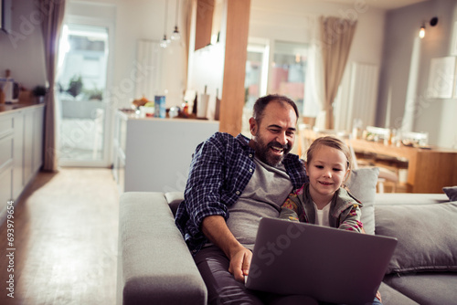 Happy little girl sitting on couch with father using laptop