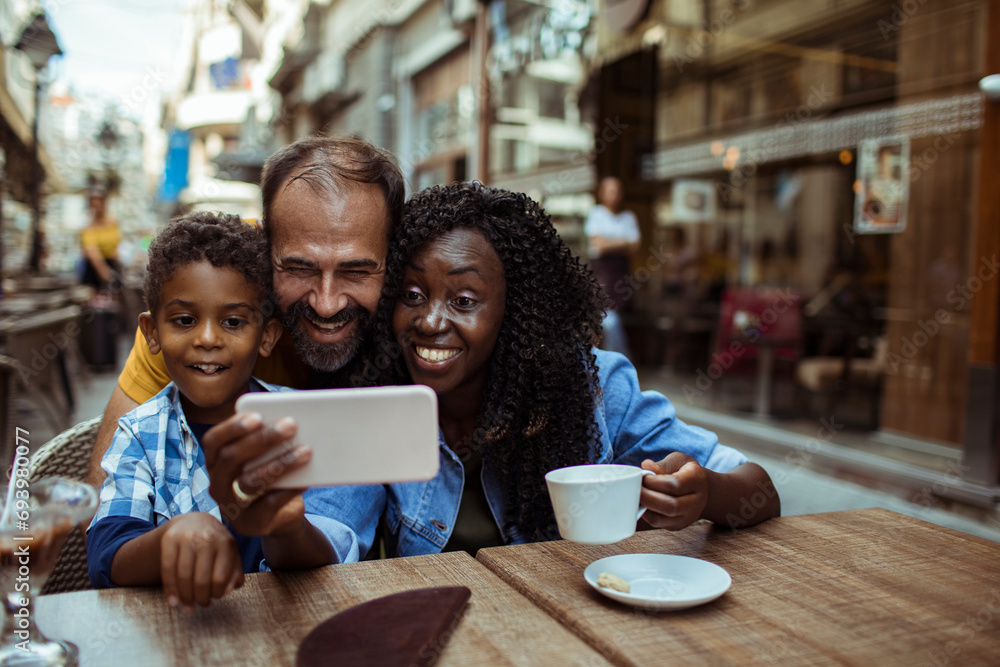 Happy multiracial couple taking selfie with little son sitting in outdoor cafe Happy multiracial couple taking selfie with little son sitting in outdoor cafe