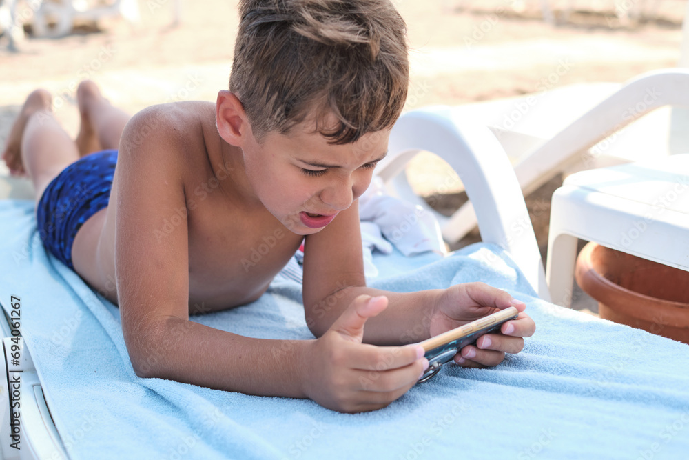 A boy lounges absorbed in his phone, with a sandy beach backdrop. Emphasizes the digital invasion into traditional outdoor settings.