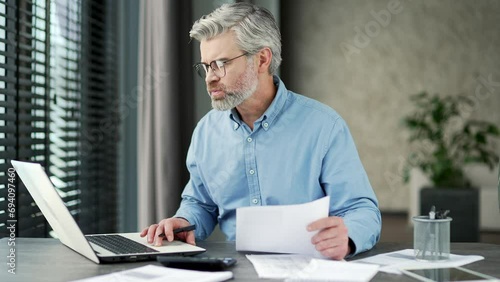 Confused puzzled mature gray haired businessman having difficulty with paper work sitting in business office. Frustrated financier uses computer reviewing documents, unhappy with bad financial results photo