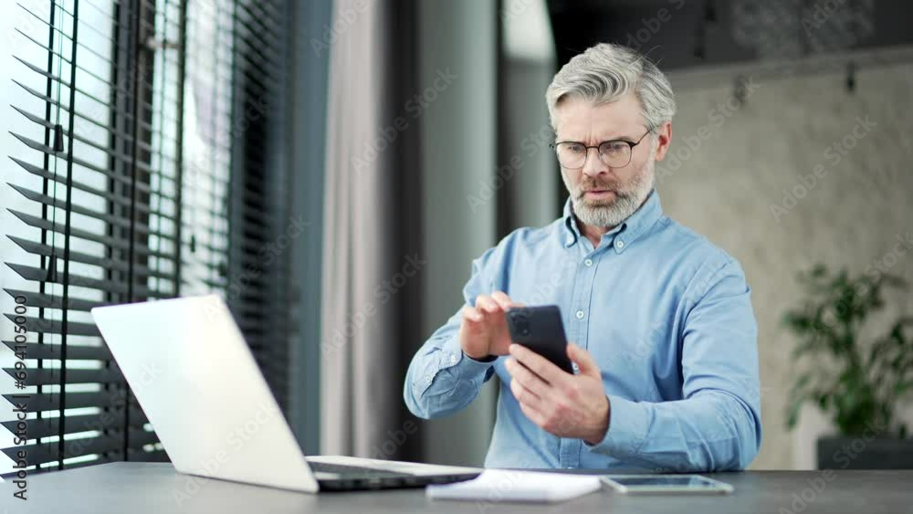 Frustrated stressed mature gray haired bearded businessman owner reads bad news on smartphone sitting at workplace in business office. Disappointed investor is upset reviewing negative information Frustrated stressed mature gray haired bearded businessman owner reads bad news on smartphone sitting at workplace in business office. Disappointed investor is upset reviewing negative information
