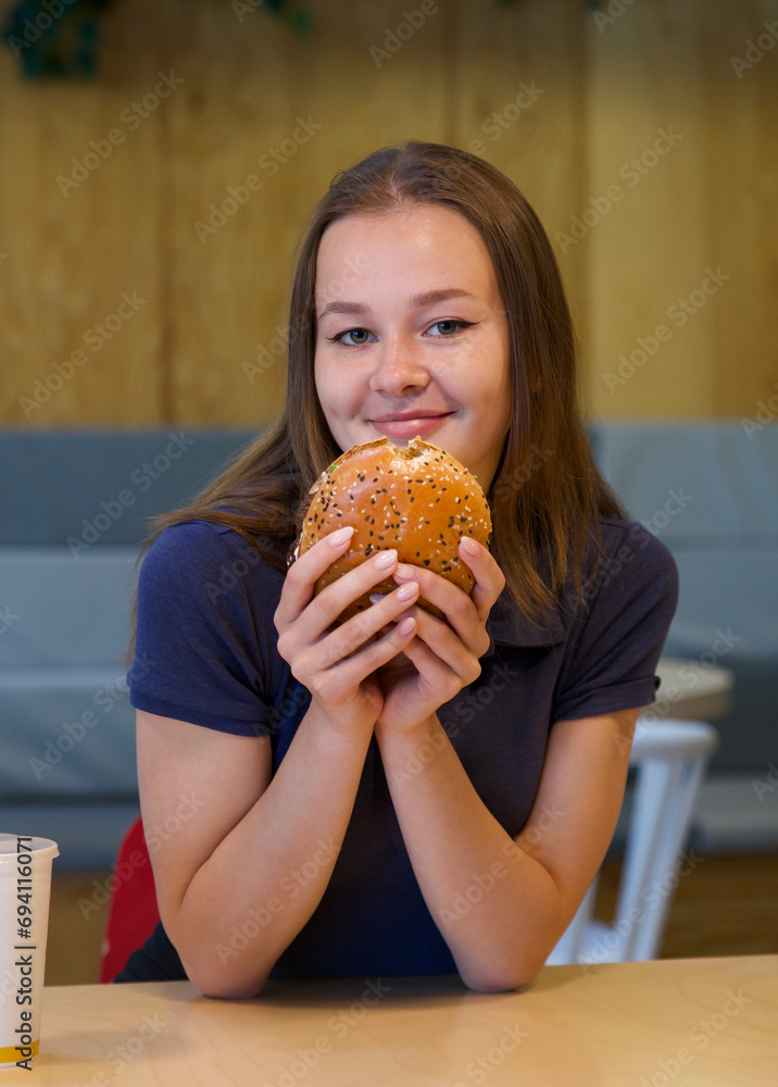 Portrait of young happy woman or beautiful teenager girl eating fast junk food, tasty burger and drink soda in a restaurant or cafe, enjoy meal. 