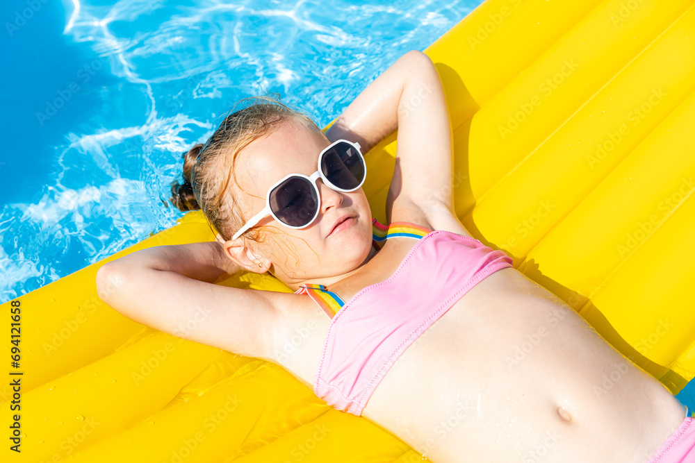 Cute little girl lying on inflatable mattress in swimming pool with blue water on warm summer day on tropical vacations. Summertime activities concept. Cute little girl sunbathing on air mattress - obrazy, fototapety, plakaty 