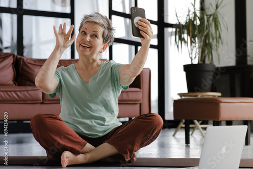 A middle aged lady practices yoga in the large hall of the house. A smiling woman sits on a mat on the floor in the lotus position, has a break time from training, uses a smartphone for video photo