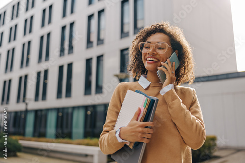 Smiling woman manager is talking phone while standing with laptop on modern building background photo