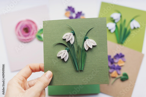 Hand holding quilling card with Snowdrops flowers. woman making greeting cards. Hand made of paper quilling technique. Handicraft at home. Hobby, home office. photo