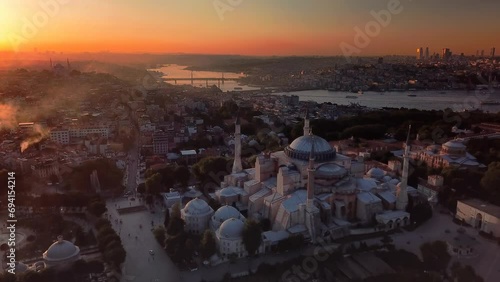 Dome of Hagia Sophia and the Goldenhorn shines in distance at sunset over historic old Istanbul. Chimney smokes from houses rises to sky against bright lights of the sun on a cold winter day
 photo