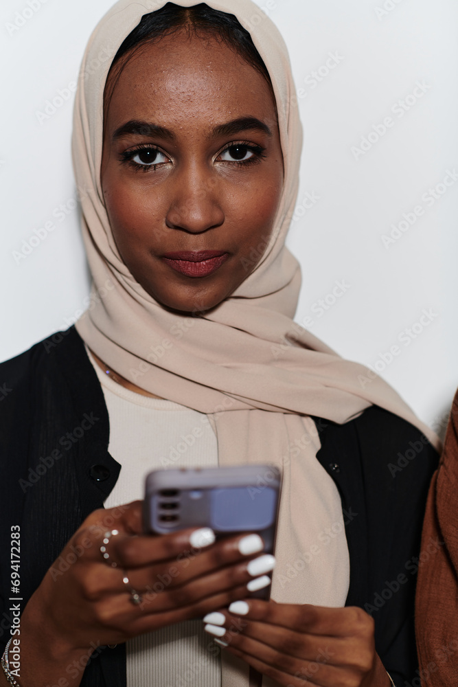 An elegant Arab woman, adorned in a hijab, engages with modernity as she uses a smartphone, the juxtaposition of traditional attire against contemporary technology captured in the isolated setting