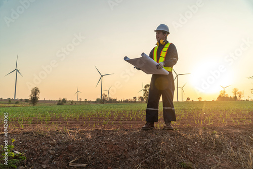 Male engineer in uniform wearing helmet stands and monitors wind turbine station operation holding blueprints using renewable energy until sunset photo