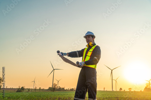 Male engineer in uniform wearing helmet sits and monitors wind turbine station operation holding blueprints checking work using renewable energy until sunset