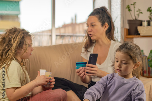a mother with a bank card and a smartphone explains to her little children with dreadlocks about finances at home, financial technology, copy space