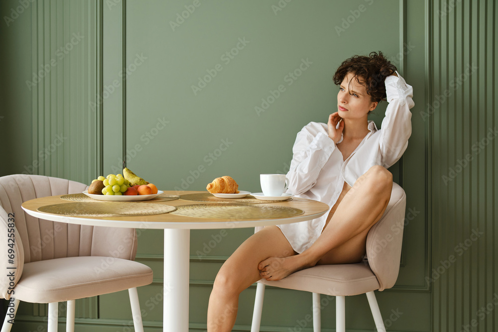 Pensive woman in white shirt sits in her modern apartment and having healthy breakfast