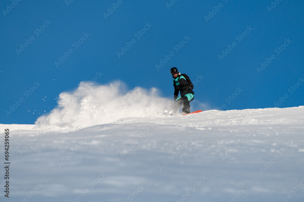 One-armed male snowboarder in a black and blue suit and protective helmet maneuvers down the slope