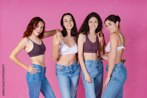 Four women in denim jeans radiate confidence and body positivity, standing together against a pink tone