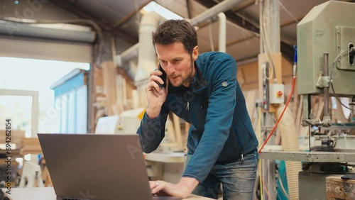 Male carpenter working in woodwork workshop talking on mobile phone whilst using laptop - shot in slow motion