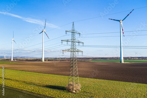 Electricyl pylon with cables on an agricultural field surrounded by windmills for wind power production