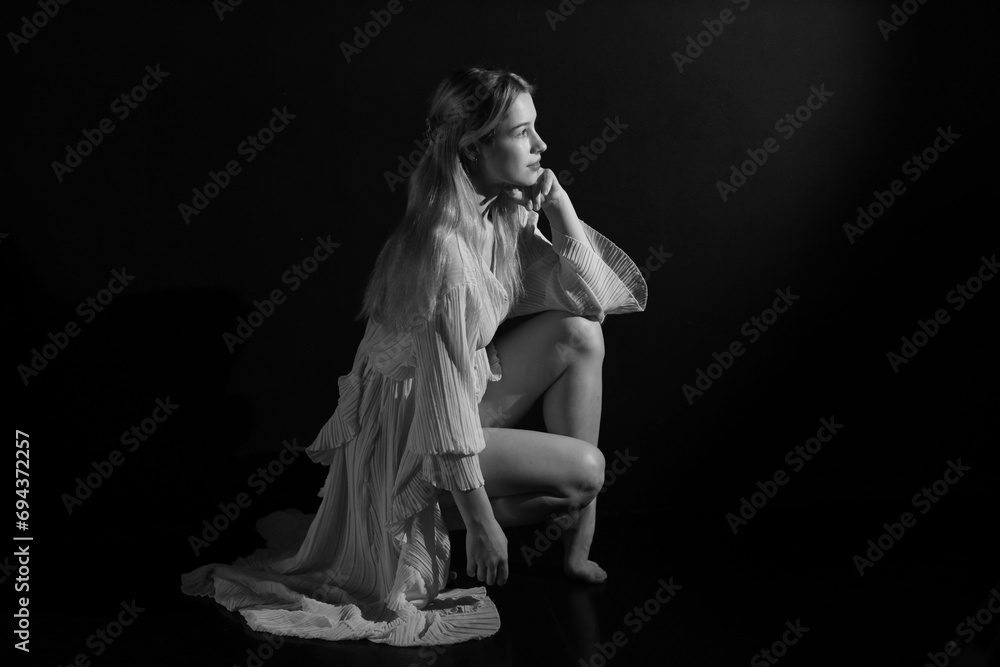 Low key black and white portrait of young pretty woman in vintage white dress sitting on the floor