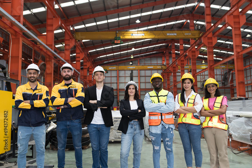 Group of male and female factory workers standing together with crossed arms and smiling in industry factory, wearing safety uniform and helmet. Factory workers working in factory