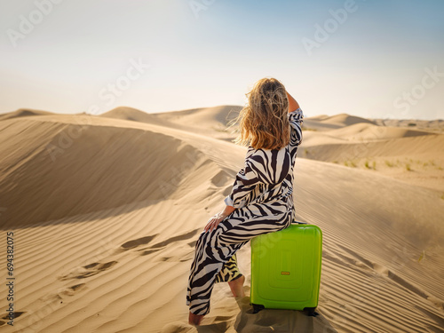 Arabian woman in stylish zebra suit with bright light green or yellow suitcase in sands. Concept and idea of travel to United Arab Emirates, sand dunes in desert of Dubai at sunset. photo